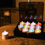 Een set van 12 RGB LED-Waxine/theelichtjes oplaadbaar waterdicht kaarsen met afstandsbediening staan in een open zwarte kist op een ronde houten tafel, één buiten de kist geplaatst; een moderne woonkamer is op de achtergrond.