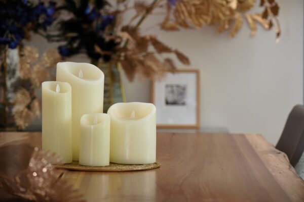 De LED wax kaarsen set 4 kaarsen warm wit oplaadbaar DORSETTO staat samen op een houten tafel, omringd door herfstbladeren met een vage ingelijste foto op de achtergrond.