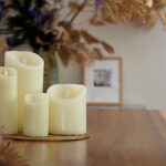 Vier Oplaadbare LED waxinelichtjes met bewegende vlam D 7,5 X 10 CM - warm wit in ivoor zijn gerangschikt op een houten tafel met gouden accenten. De onscherpe achtergrond is voorzien van gedroogd gebladerte en ingelijste kunst voor een elegante, gezellige sfeer.