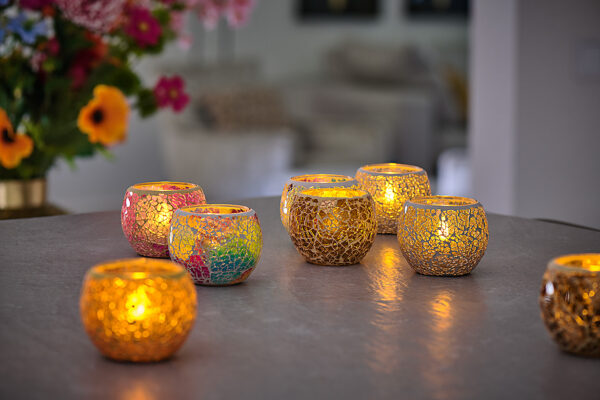 Zes decoratieve mozaïek kandelaars met LED- waxine/theelichtjes waterdicht oplaadbaar (warm wit, 175 uur brandtijd, 12 stuks, met afstandsbediening) staan op een tafel; drie zijn goudkleurig en drie zijn veelkleurig. Bloemen en neutrale meubels vervagen op de achtergrond.