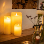 Drie Oplaadbare LED wax kaarsen met bewegende vlam (D 7,5 x 12,5 cm, warm wit) staan te gloeien op een houten oppervlak naast een muur, met vage bloemenvazen ervoor, en creëren een gezellige en uitnodigende sfeer.