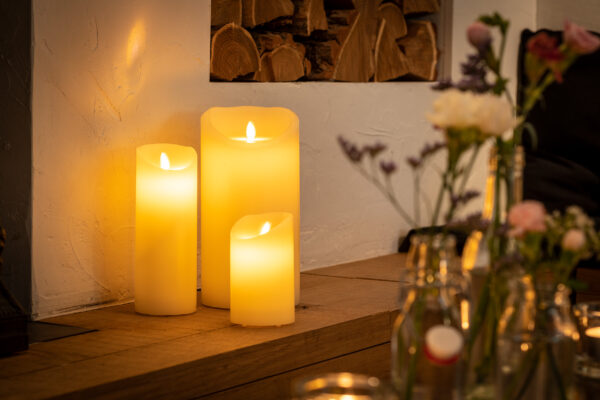 Drie Oplaadbare LED wax kaarsen met bewegende vlam (D 7,5 x 12,5 cm, warm wit) staan te gloeien op een houten oppervlak naast een muur, met vage bloemenvazen ervoor, en creëren een gezellige en uitnodigende sfeer.