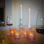 Een close-up van een gedekte tafel met verschillende brandende kaarsen in geribbelde glazen houders, twee Oplaadbare LED dinerkaarsen 270mm (130 uur, warm wit, 12 stuks met afstandsbediening), een glas rode wijn, een kruik en groene druiven voor een gezellige sfeer.