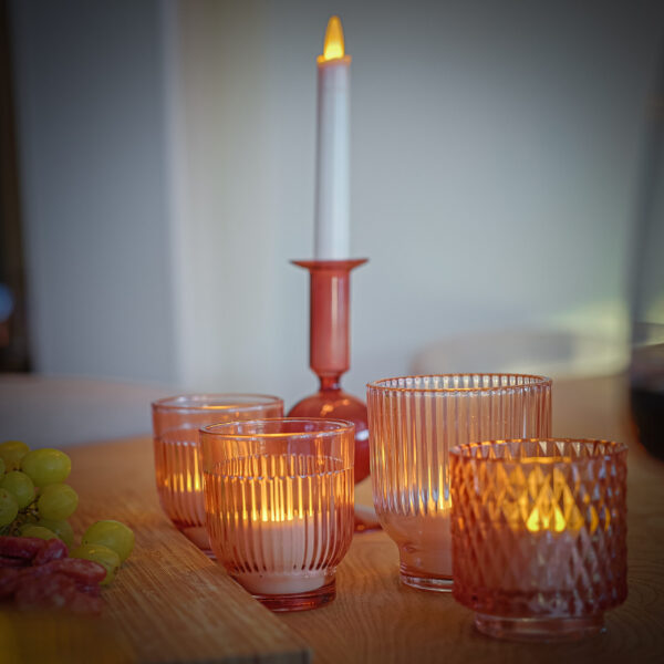 Roze glazen kandelaars en een Dinerkaars 197mm LED DORSETTO oplaadbaar 130 uur warm wit (12 stuks, met afstandsbediening) staan op een houten tafel met groene druiven en gesneden gerookt vlees aan de linkerkant, waardoor een zacht verlicht, gezellig tafereel ontstaat.