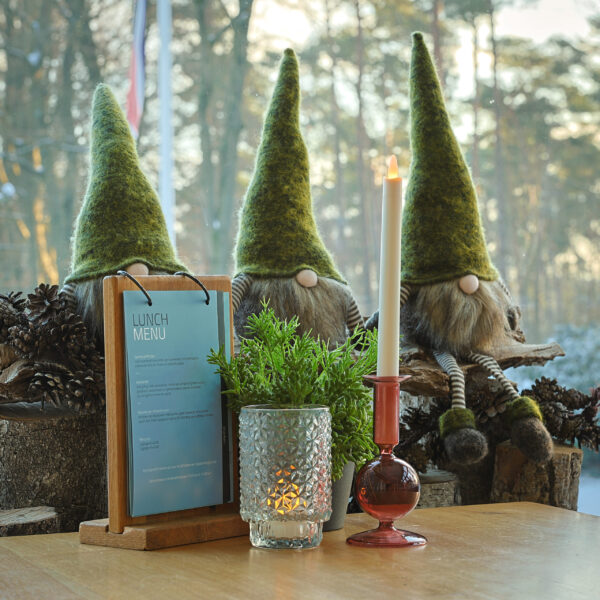 Een gezellige tafel met een lunchkaart, glas met structuur, potplant, LED dinerkaars 270mm oplaadbaar 130 uur warm met 6 stuks met afstandsbediening, en drie decoratieve kabouters met lange groene hoeden bij een besneeuwd bosraam.