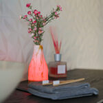 Een gloeiende Tafellamp DORSETTO oplaadbaar 105 uur (set van 2, waterdicht, marmer goud, met afstandsbediening) staat naast een vaas met roze bloemen op een donker oppervlak in de buurt van bamboe tandenborstels en handdoeken, met een rietverdamper op de achtergrond.