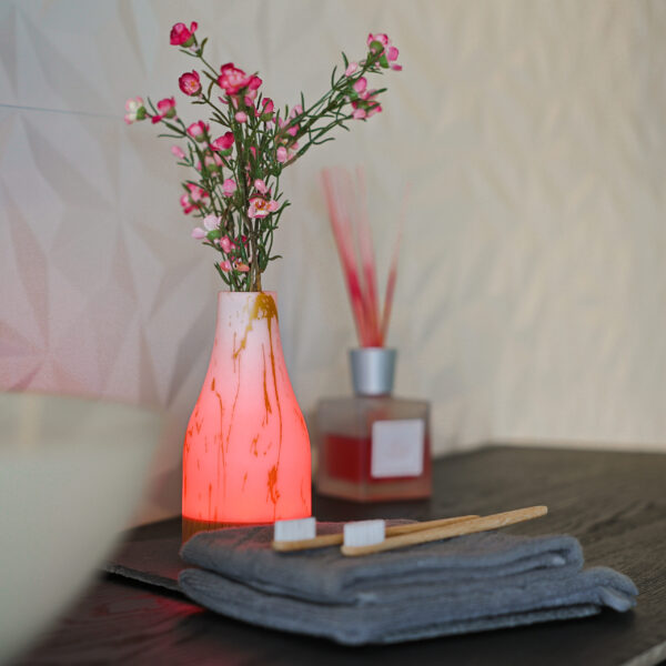 Een gloeiende Tafellamp DORSETTO oplaadbaar 105 uur (set van 2, waterdicht, marmer goud, met afstandsbediening) staat naast een vaas met roze bloemen op een donker oppervlak in de buurt van bamboe tandenborstels en handdoeken, met een rietverdamper op de achtergrond.