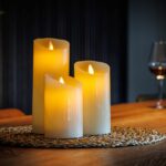 Drie oplaadbare LED wax kaarsen van 3 stuks DORSETTO in verschillende hoogtes verspreiden een warme gloed op een gevlochten mat op een houten tafel, terwijl op de achtergrond een wijnfles en gevuld glas zorgen voor een gezellige sfeer.