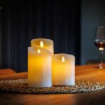 De LED wax kaarsen set 3 kaarsen DORSETTO met een warme gele gloed staat op een geweven placemat op een houten tafel. Op de zacht vervaagde achtergrond staan een glas rode wijn en een fles voor een gezellige, ontspannen sfeer.