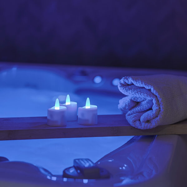 Een ontspannen spascène met oplaadbare, waterdichte RGB LED-Waxine/theelichtjes (12 stuks) met afstandsbediening op een houten plank bij een opgerolde handdoek, zacht oplichtend in blauw licht naast een bubbelend bubbelbad.