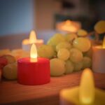 Een close-up van een rood Oplaadbaar LED-kaarsje (set van 12, waterdicht, met afstandsbediening en timerfunctie) op een houten oppervlak met kunstkaarsen en groene druiven, waardoor een gezellige sfeer ontstaat met een zacht wazige achtergrond.
