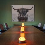 Een houten tafel met drie gloeiende Tafellamp DORSETTO oplaadbaar 105 uur (waterdicht, marmer groen houtlook, met afstandsbediening) lampen wordt omringd door stoelen. Daarachter zorgt een zwart-wit highland koeienzeil voor een rustige, moderne vibe.