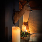 Een warm verlicht interieur heeft een gemonteerde hertenkop boven een schouw, versierd met de Oplaadbare LED wax kaars D 7,5 X 15 CM - warm wit en een groen middenstuk. Zachte schaduwen en gezellige verlichting maken de ruimte uitnodigend.
