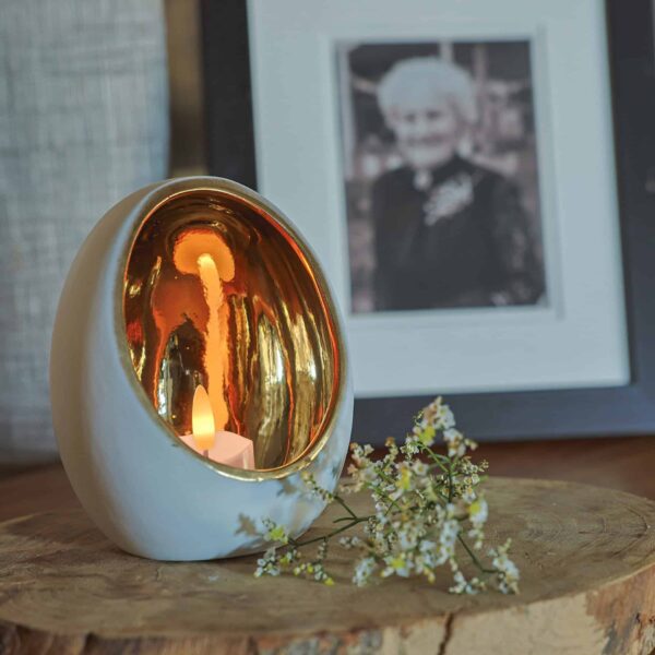 Een 3D oplaadbare LED-Waxine/theelichtje uit een set van 12 met timerfunctie zit te gloeien in een gouden eivormige houder op hout naast witte bloemen, voor een ingelijst zwart-wit portret van een oudere vrouw.