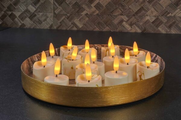 Een rond gouden dienblad toont 12 3D oplaadbare LED-Waxine/theelichtjes (175 uur, met afstandsbediening, timerfunctie), hun kunstmatige vlammen dicht bij elkaar geplaatst voor een warme, gezellige sfeer tegen donkere tegels.