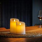 Twee LED kleine wax kaarsen uit de set van 2 kaarsen DORSETTO staan op een placemat op een houten tafel met op de achtergrond een glas rode wijn en fles voor een warme, gezellige sfeer.