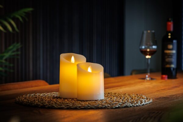 Twee LED kleine wax kaarsen uit de set van 2 kaarsen DORSETTO staan op een placemat op een houten tafel met op de achtergrond een glas rode wijn en fles voor een warme, gezellige sfeer.