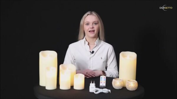 Een vrouw in een wit shirt zit aan een zwarte tafel met de DORSETTO LED waxinelichtjesset 2 maten warm wit oplaadbaar, twee glitterende waxinelichthouders, een afstandsbediening en een oplaadkabel, tegen een zwarte achtergrond met het Dorsetto-logo.