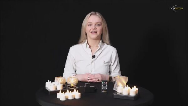 Een vrouw met lang blond haar in een wit shirt zit aan een ronde zwarte tafel met brandende kaarsen en het Tafellampje DORSETTO oplaadbaar 105 uur (waterdicht, ovaal, marmer groen houtlook grijs goud, met afstandsbediening), tegen een zwarte achtergrond.