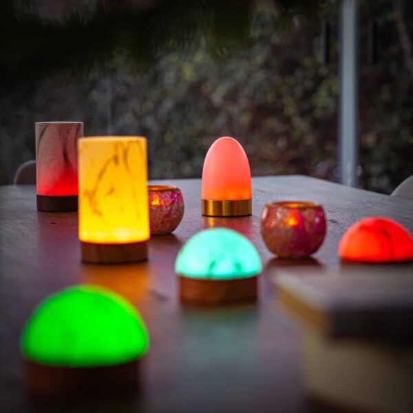 Op een houten tafel staat de Tafellamp DORSETTO oplaadbaar (105 uur, waterdicht, ovaal, marmer wit-grijs houtlook, met afstandsbediening) tussen andere kleurrijke lampen, die een gezellige sfeer creëren. Onscherpe bomen vullen de achtergrond.