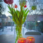 Een heldere glazen pot met roze en witte tulpen staat op een houten tafel naast twee brandende Waxinelichthouder 8,5 CM Dorsetto mozaïek rozenkandelaars. Op de achtergrond staat een besneeuwde tuin met charmante huisjes en bomen.