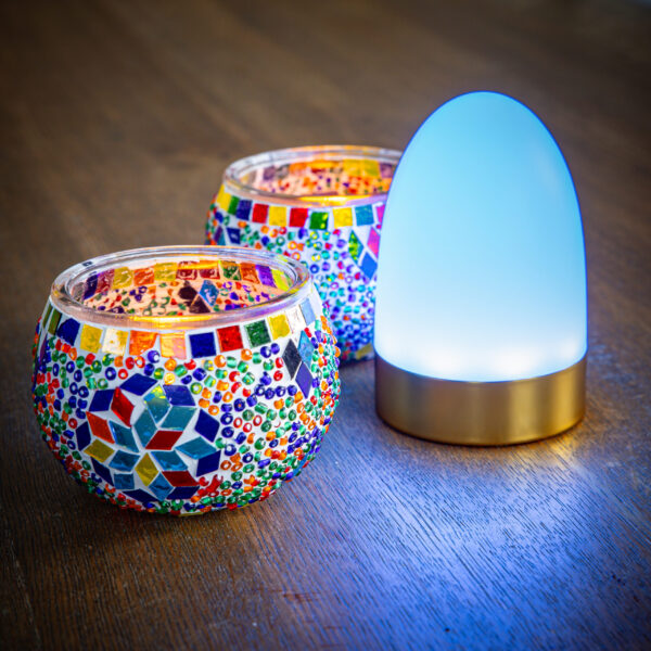 Twee ronde, kleurrijke kandelaars in mozaïek staan naast een Tafellamp DORSETTO oplaadbaar 105 uur, een waterdichte, ovale LED-lamp in witgoud met afstandsbediening, op een houten oppervlak.