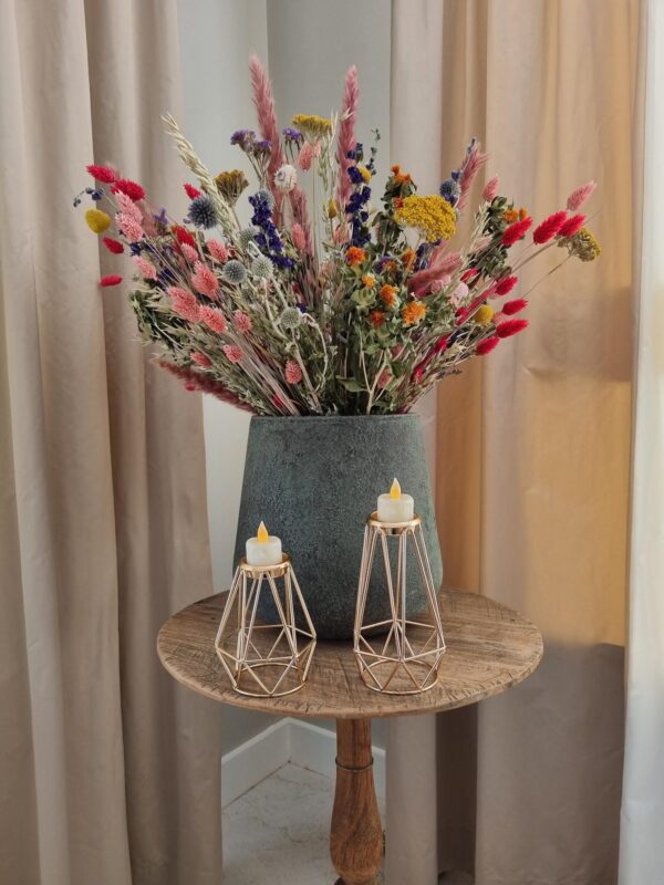 Op een rustieke houten ronde tafel staat een grote vaas met kleurrijke droogbloemen en een Waxinelichthouderset DORSETTO metaal rosé goudig per 2 stuks met kleine brandende kaarsjes, tegen beige gordijnen.