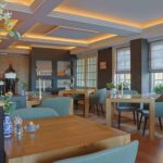 Een gezellig, modern restaurant met houten tafels, zachtgroene stoelen, warme verlichting en grote ramen heeft een elegante inrichting met vazen, planten en LED-Teelicht wiederaufladbar mit Fernbedienung für 175 Stunden Brenndauer.
